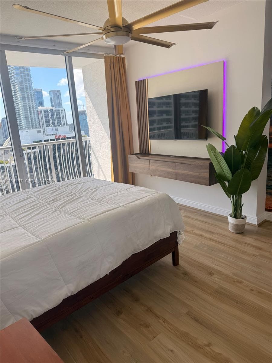 Floor-to-ceiling windows with balcony access, wood-finish flooring, a contemporary floating wood-finish media console, custom accent paneling with integrated lighting, and a modern brushed-gold ceiling fan
