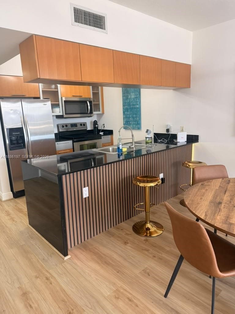 Kitchen featuring a central island with a slatted wood-style base and stone-surface countertop, stainless steel appliances, upper wood cabinetry, and wood-finish flooring