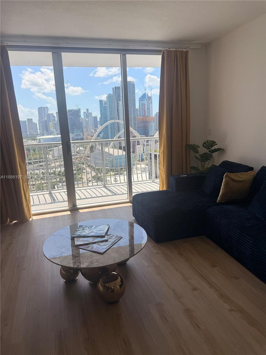 Floor-to-ceiling windows, wood-finish flooring, and a private balcony featuring a metal railing and metropolitan cityscape backdrop