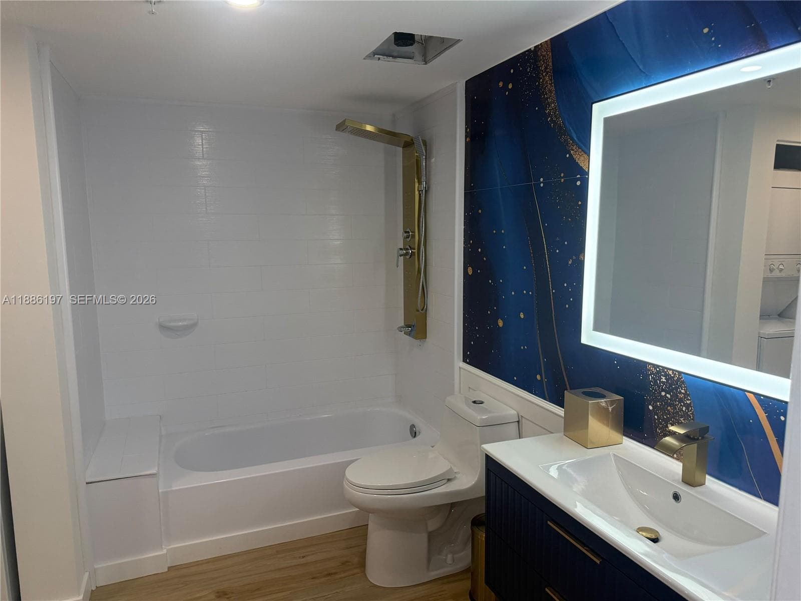 Bathroom featuring a white subway tile tub surround, gold-finish shower panel, wood-finish flooring, dark blue cabinetry with gold hardware, and a back-lit rectangular mirror