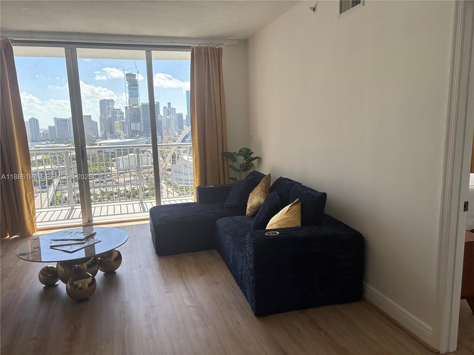 Floor-to-ceiling glass sliding doors, private balcony with metal railing, light-tone wood-finish flooring, and expansive urban skyline backdrop