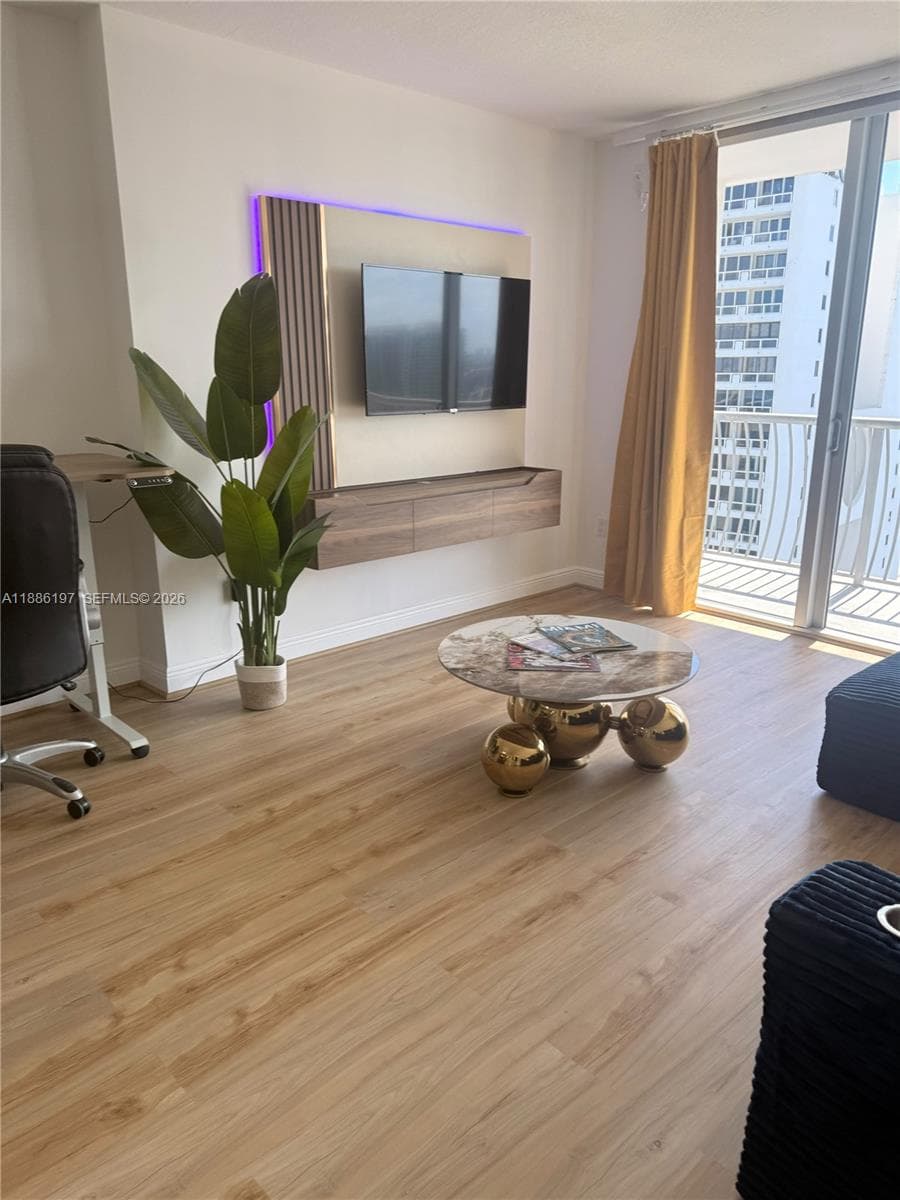 Wood-finish flooring, custom wall-mounted media console with integrated backlighting, and floor-to-ceiling glass sliding doors opening to a private balcony