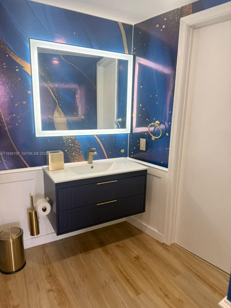 Wall-mounted vanity featuring dual drawers, integrated sink, gold-toned hardware, and an LED-backlit mirror set against decorative wall treatments with white wainscoting and wood-finish flooring