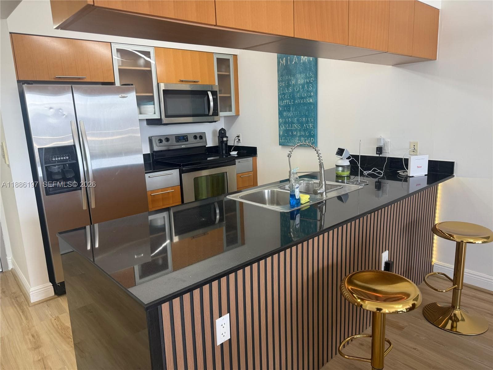 Kitchen featuring wood-finish cabinetry, black stone countertops, stainless steel appliances, and a slatted wood-paneled kitchen island with an undermount sink and pull-down faucet
