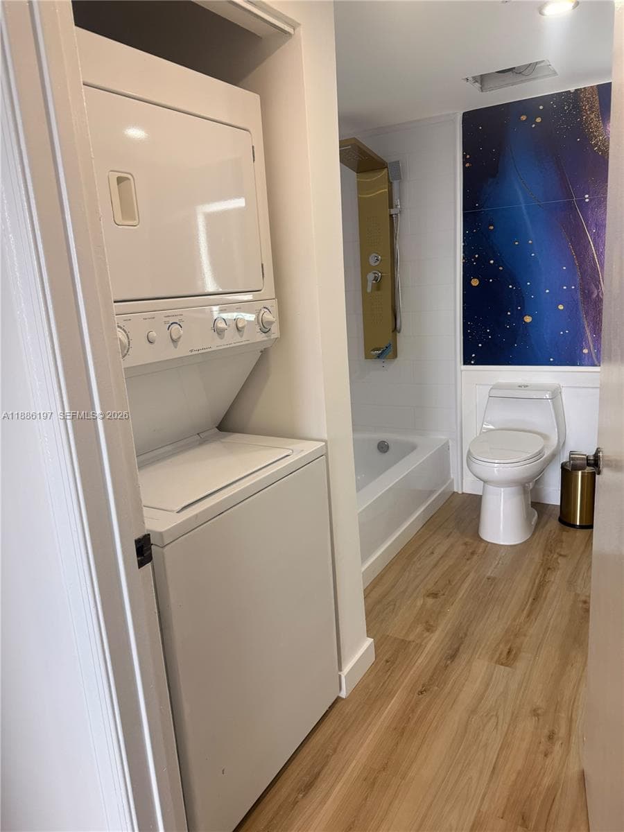Stacked laundry unit, wood-finish flooring, full-size bathtub, wall-mounted shower panel, and dual-flush toilet