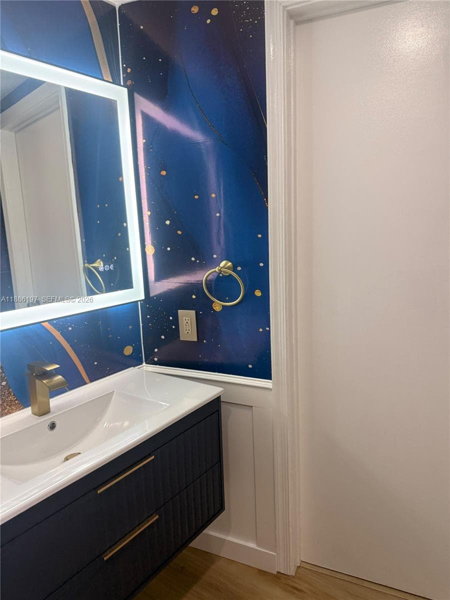 Floating vanity with a white basin, brass-finish faucet, illuminated mirror, decorative wall covering, and beadboard wainscoting