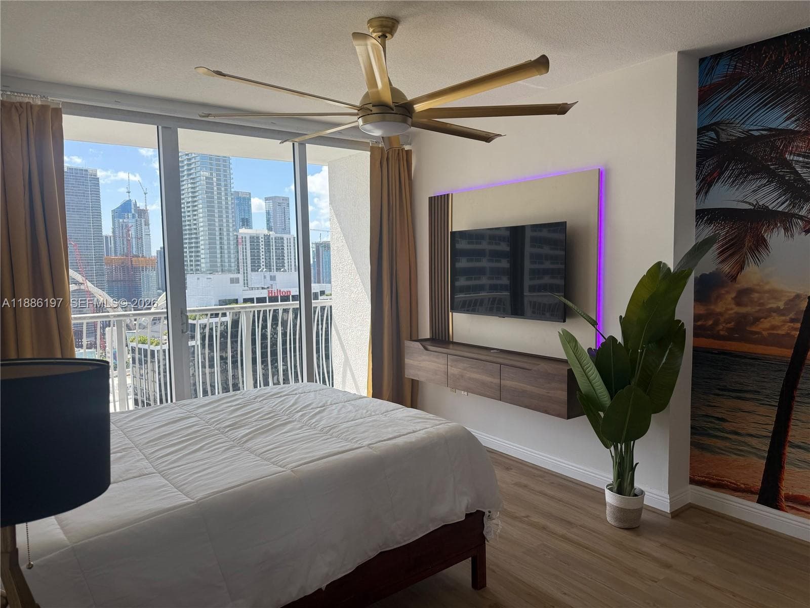 Floor-to-ceiling glass sliding doors, wall-mounted floating wood-finish media console, integrated back-lit wall panel, multi-blade ceiling fan, and wood-finish flooring