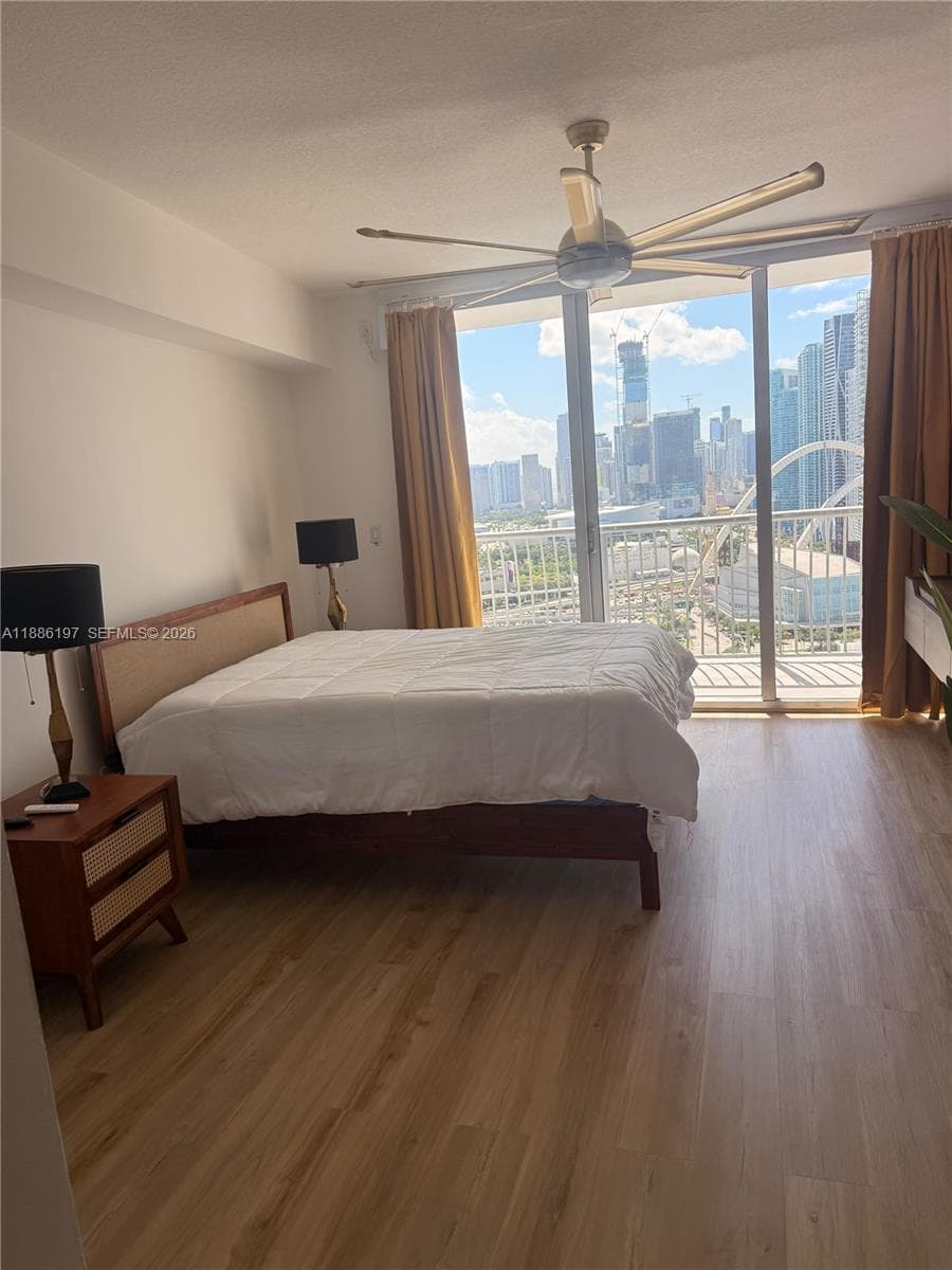 Floor-to-ceiling windows, wood-finish flooring, ceiling fan with integrated lighting, and an exterior balcony featuring metal railings with urban skyline scenery