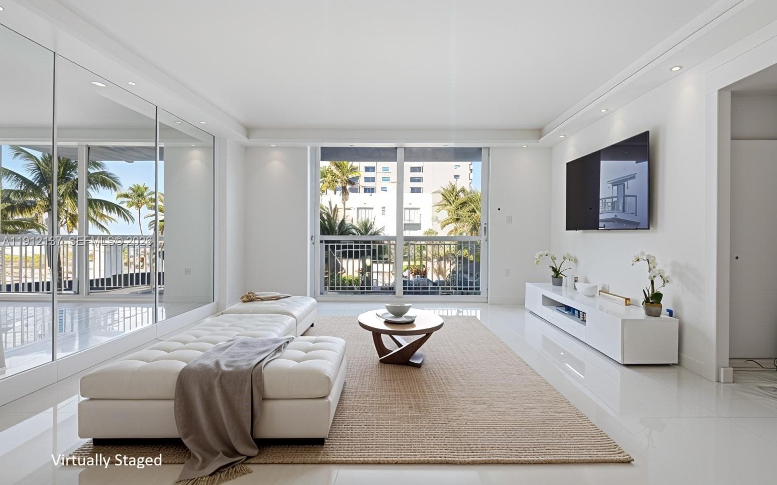 Sunlight fills the open space through floor-to-ceiling sliding doors, highlighting the gleaming floors and mirrored accent wall that enhances depth and natural light. The modern layout seamlessly connects to a private balcony with lush palm views, creating a serene indoor-outdoor flow. Clean lines, recessed lighting, and refined finishes make this residence the perfect blend of luxury and tranquility