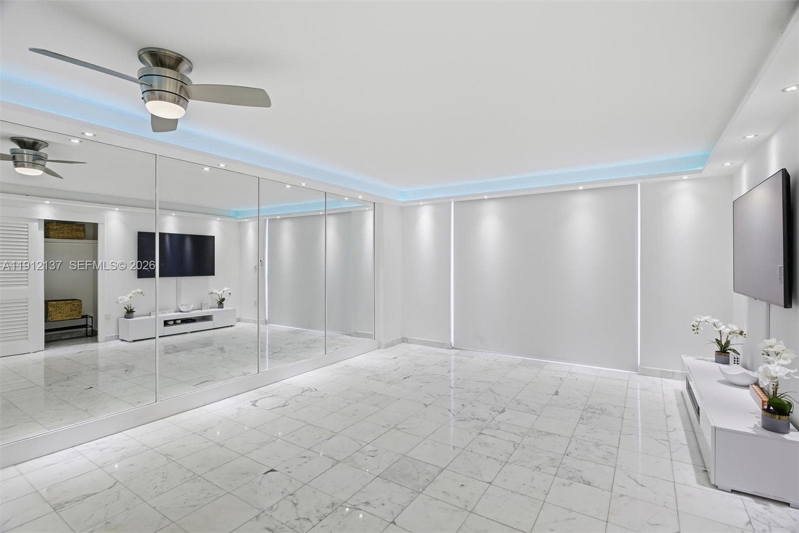 Sunlight fills the open space through floor-to-ceiling sliding doors, highlighting the gleaming floors and mirrored accent wall that enhances depth and natural light. The modern layout seamlessly connects to a private balcony with lush palm views, creating a serene indoor-outdoor flow. Clean lines, recessed lighting, and refined finishes make this residence the perfect blend of luxury and tranquility