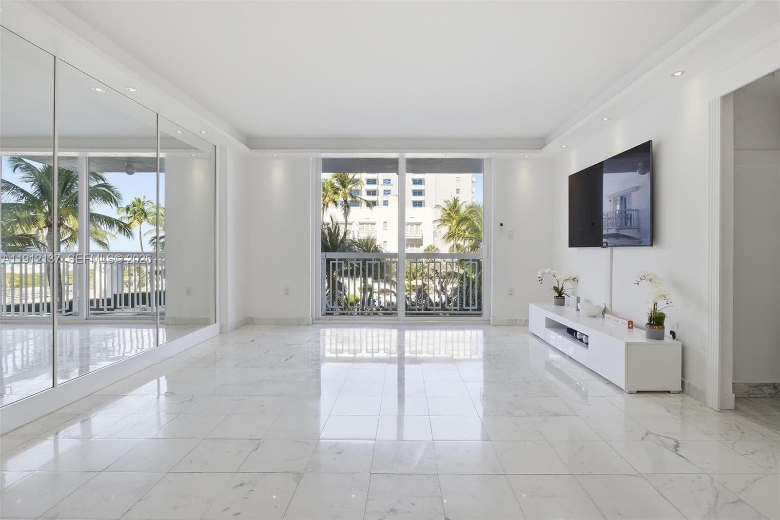 Sunlight fills the open space through floor-to-ceiling sliding doors, highlighting the gleaming floors and mirrored accent wall that enhances depth and natural light. The modern layout seamlessly connects to a private balcony with lush palm views, creating a serene indoor-outdoor flow. Clean lines, recessed lighting, and refined finishes make this residence the perfect blend of luxury and tranquility