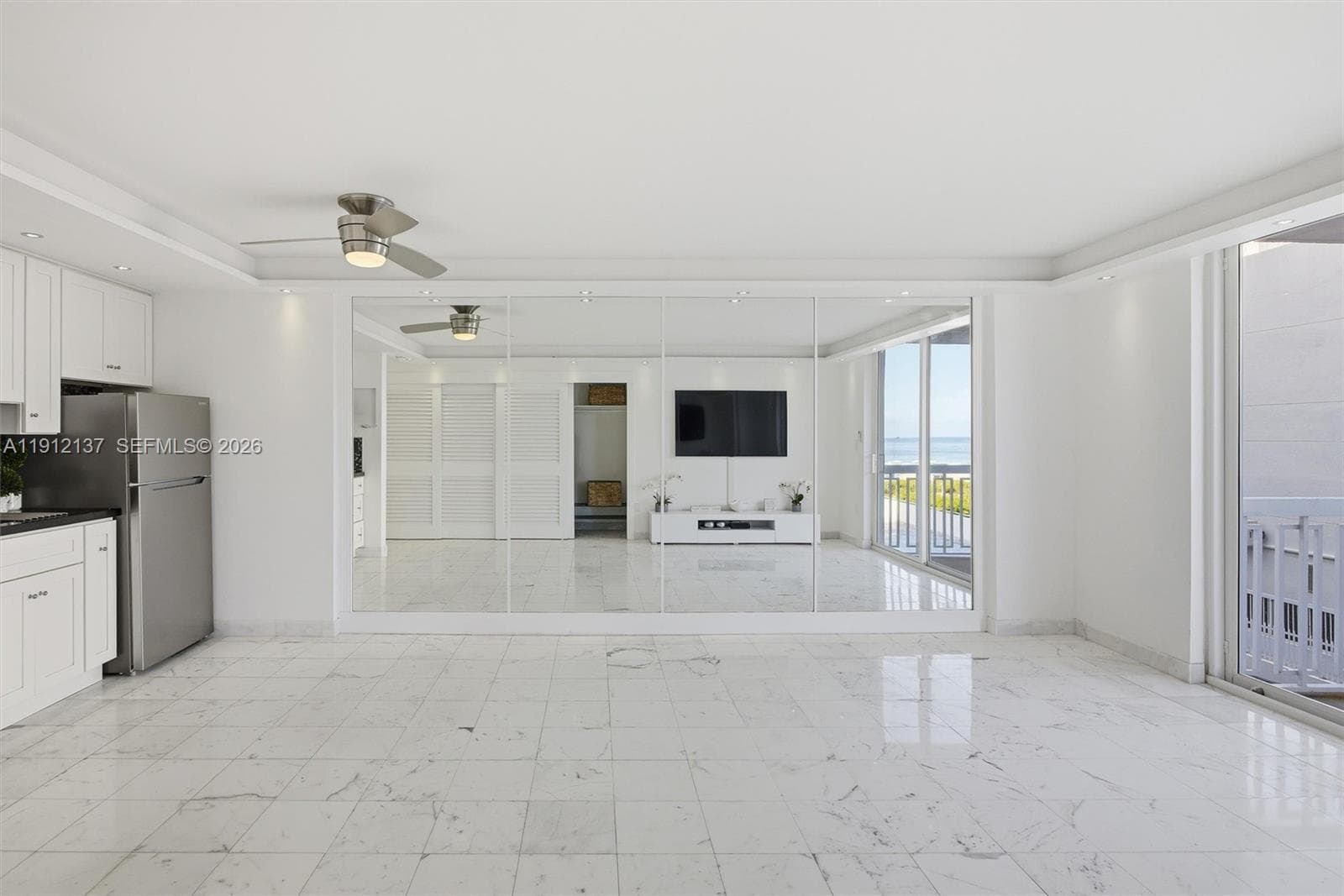 Sunlight fills the open space through floor-to-ceiling sliding doors, highlighting the gleaming floors and mirrored accent wall that enhances depth and natural light. The modern layout seamlessly connects to a private balcony with lush palm views, creating a serene indoor-outdoor flow. Clean lines, recessed lighting, and refined finishes make this residence the perfect blend of luxury and tranquility