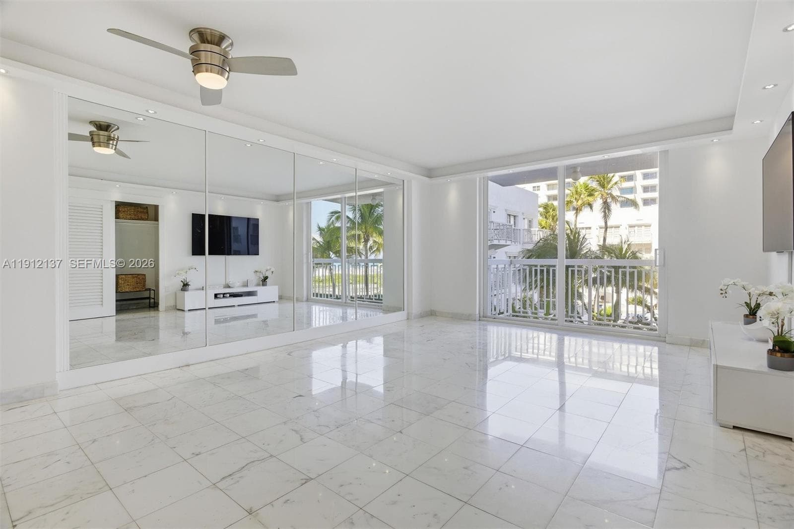 Sunlight fills the open space through floor-to-ceiling sliding doors, highlighting the gleaming floors and mirrored accent wall that enhances depth and natural light. The modern layout seamlessly connects to a private balcony with lush palm views, creating a serene indoor-outdoor flow. Clean lines, recessed lighting, and refined finishes make this residence the perfect blend of luxury and tranquility