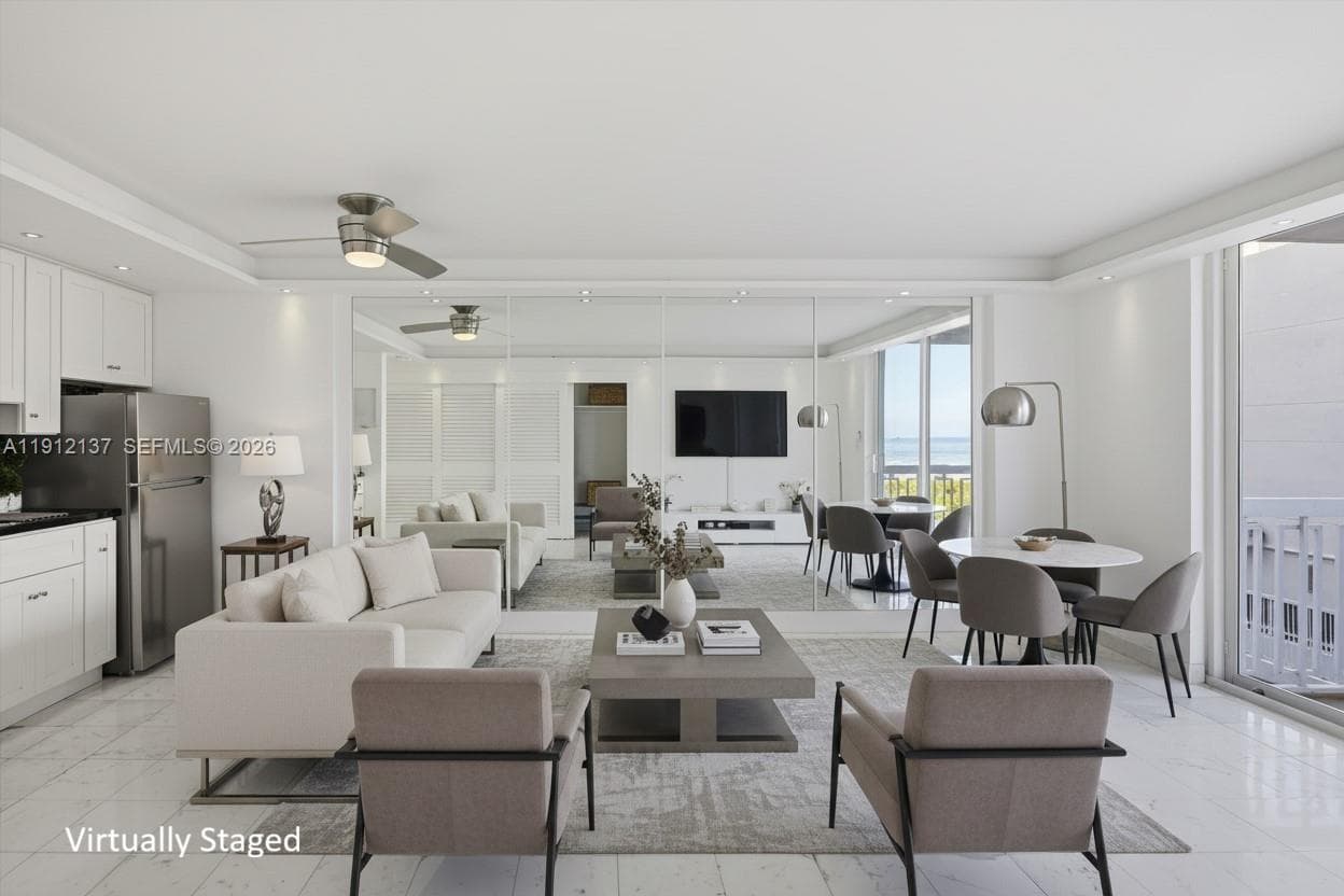 Sunlight fills the open space through floor-to-ceiling sliding doors, highlighting the gleaming floors and mirrored accent wall that enhances depth and natural light. The modern layout seamlessly connects to a private balcony with lush palm views, creating a serene indoor-outdoor flow. Clean lines, recessed lighting, and refined finishes make this residence the perfect blend of luxury and tranquility