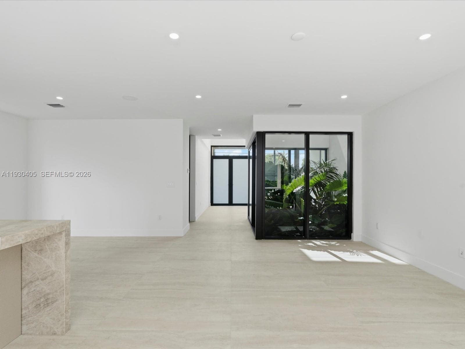 Neutral-toned stone-finish flooring, recessed ceiling lighting, interior glass-paneled garden enclosure with black framing, and stone-clad island feature