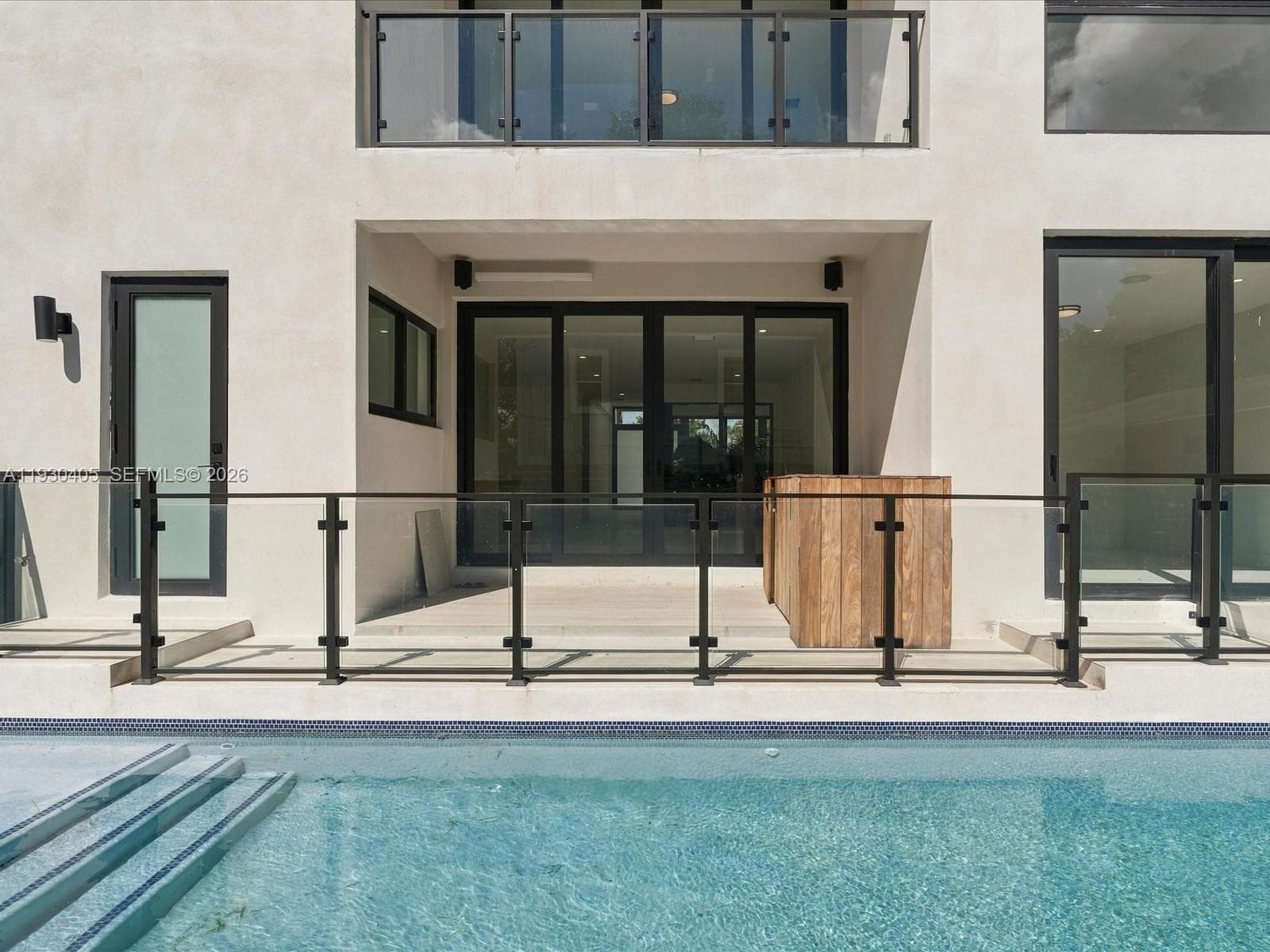 Exterior rear patio featuring smooth stucco finish, black-framed sliding glass doors, glass panel railings, and an integrated swimming pool with tiled steps