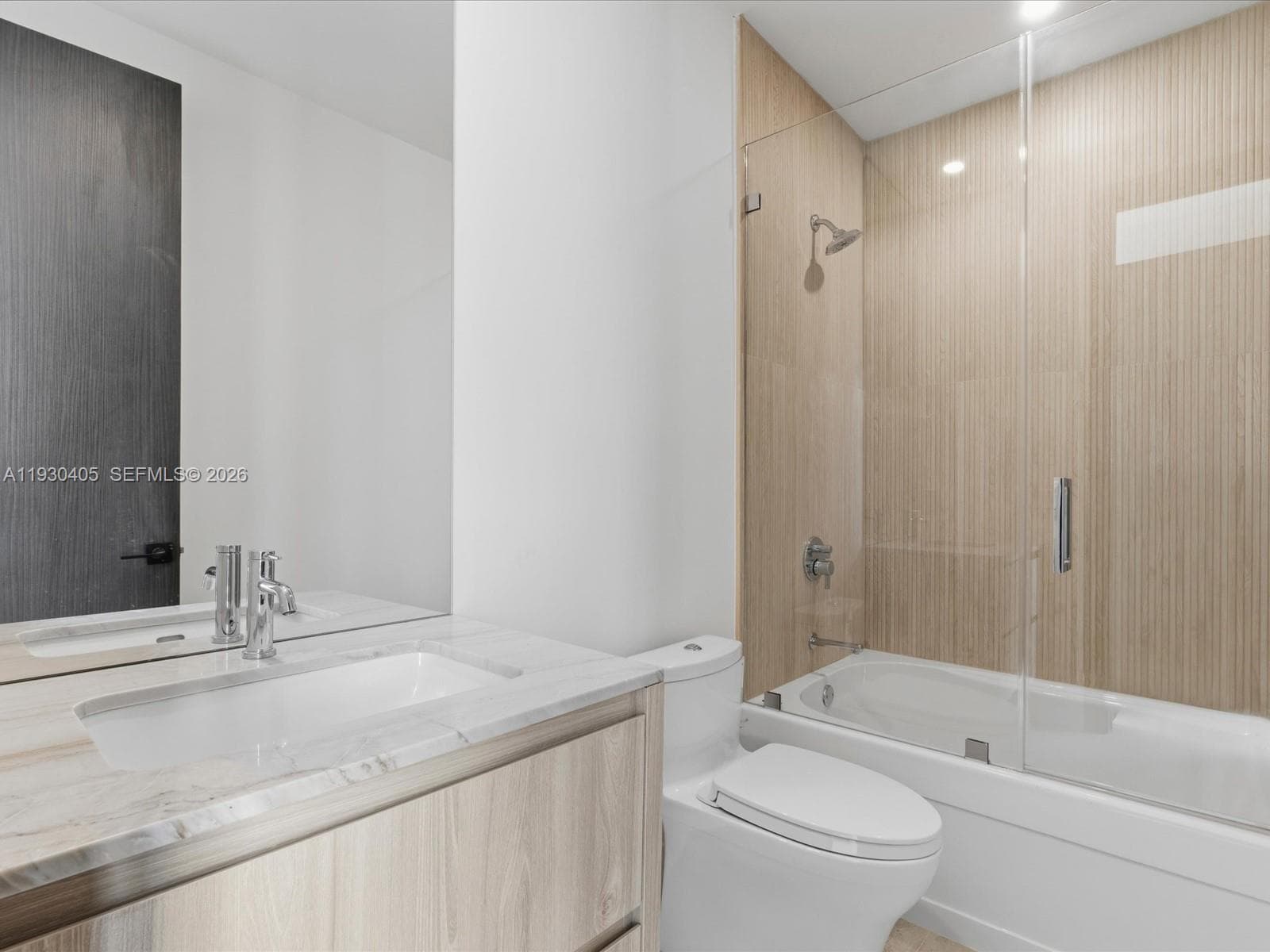 Bathroom featuring a wood-finish vanity with a stone countertop, an integrated rectangular sink, a glass-enclosed shower and tub combination, and vertical-grain wall tile