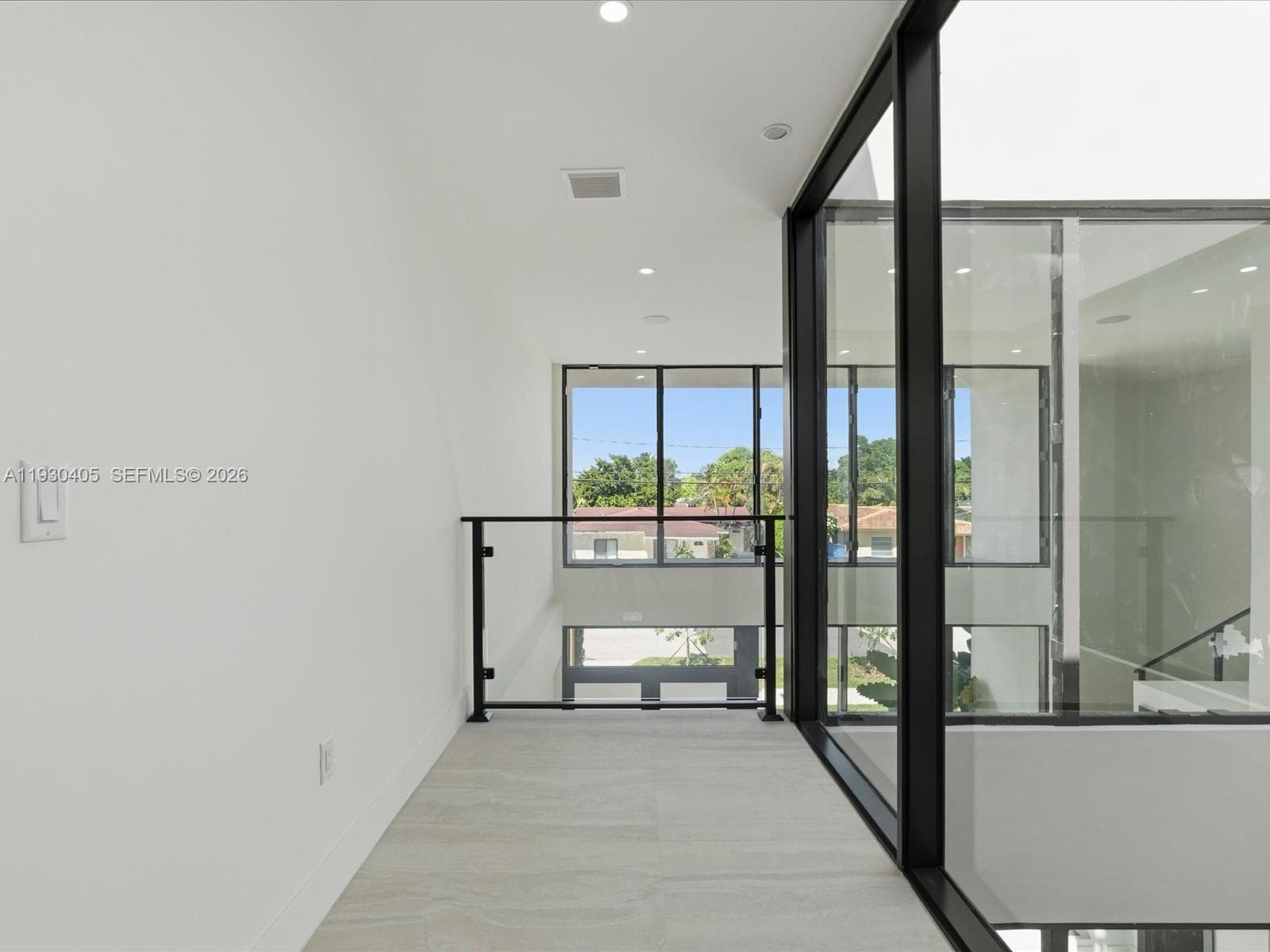 Light-filled interior featuring floor-to-ceiling black-frame windows, recessed lighting, and a glass-panel railing with black metal hardware, finished with neutral large-format tile flooring