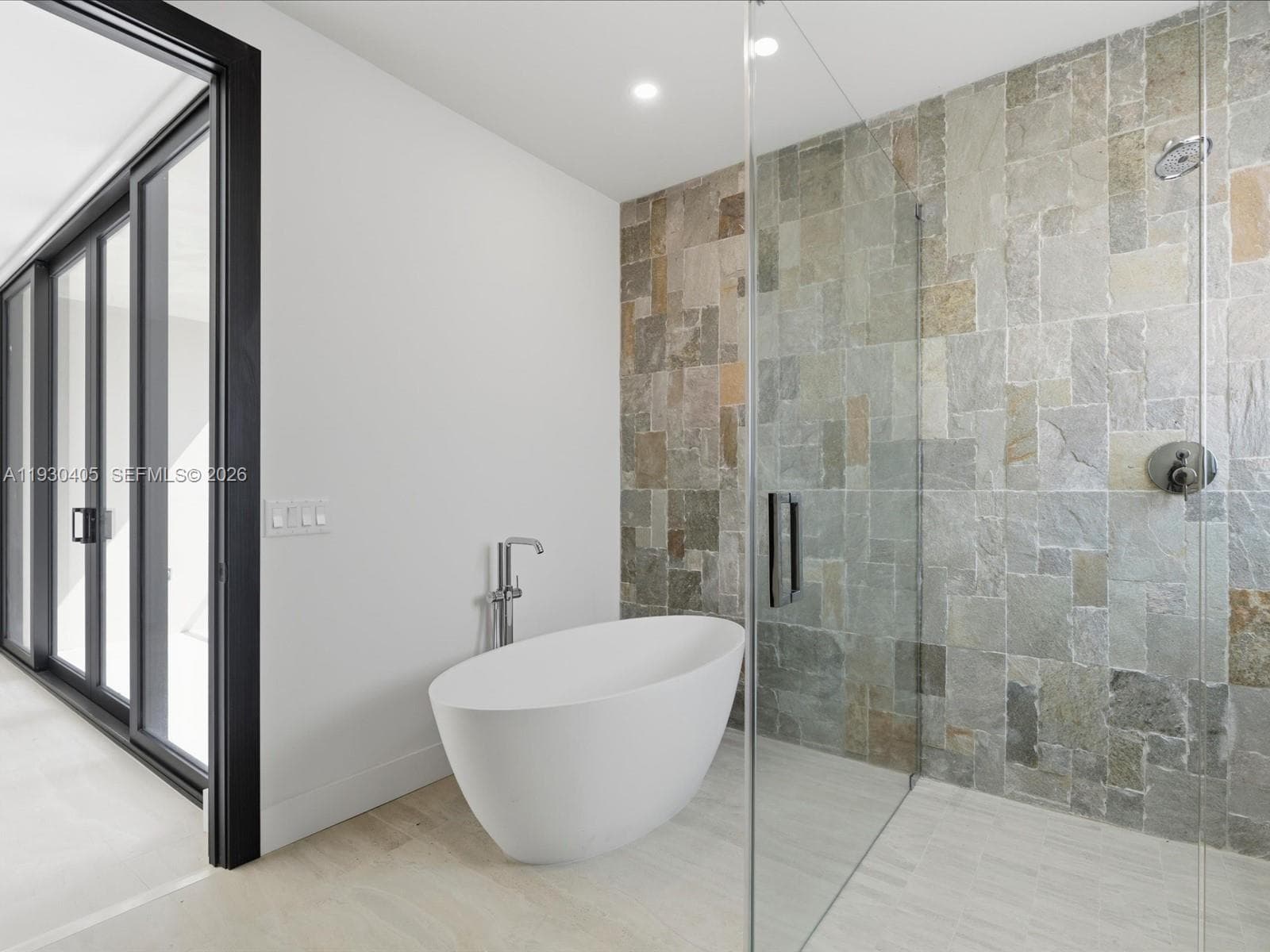 Freestanding white soaking tub with a floor-mounted faucet, glass-enclosed walk-in shower with natural stone tile wall, and sliding glass doors with black framing
