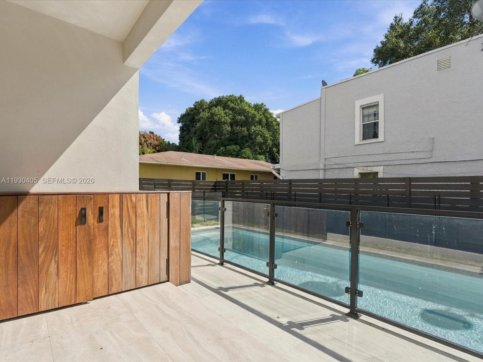 Outdoor patio featuring stone tile flooring, built-in wood-finish cabinetry, and a swimming pool enclosed by a glass-panel railing with metal frame