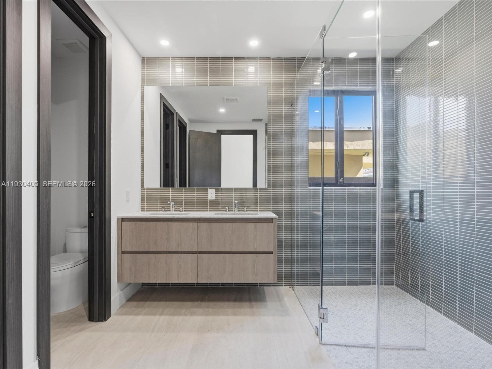Floating dual-sink vanity with light wood-finish cabinetry, countertop with integrated dual basins, full-height tile backsplash, frameless glass-enclosed shower, and recessed ceiling lighting