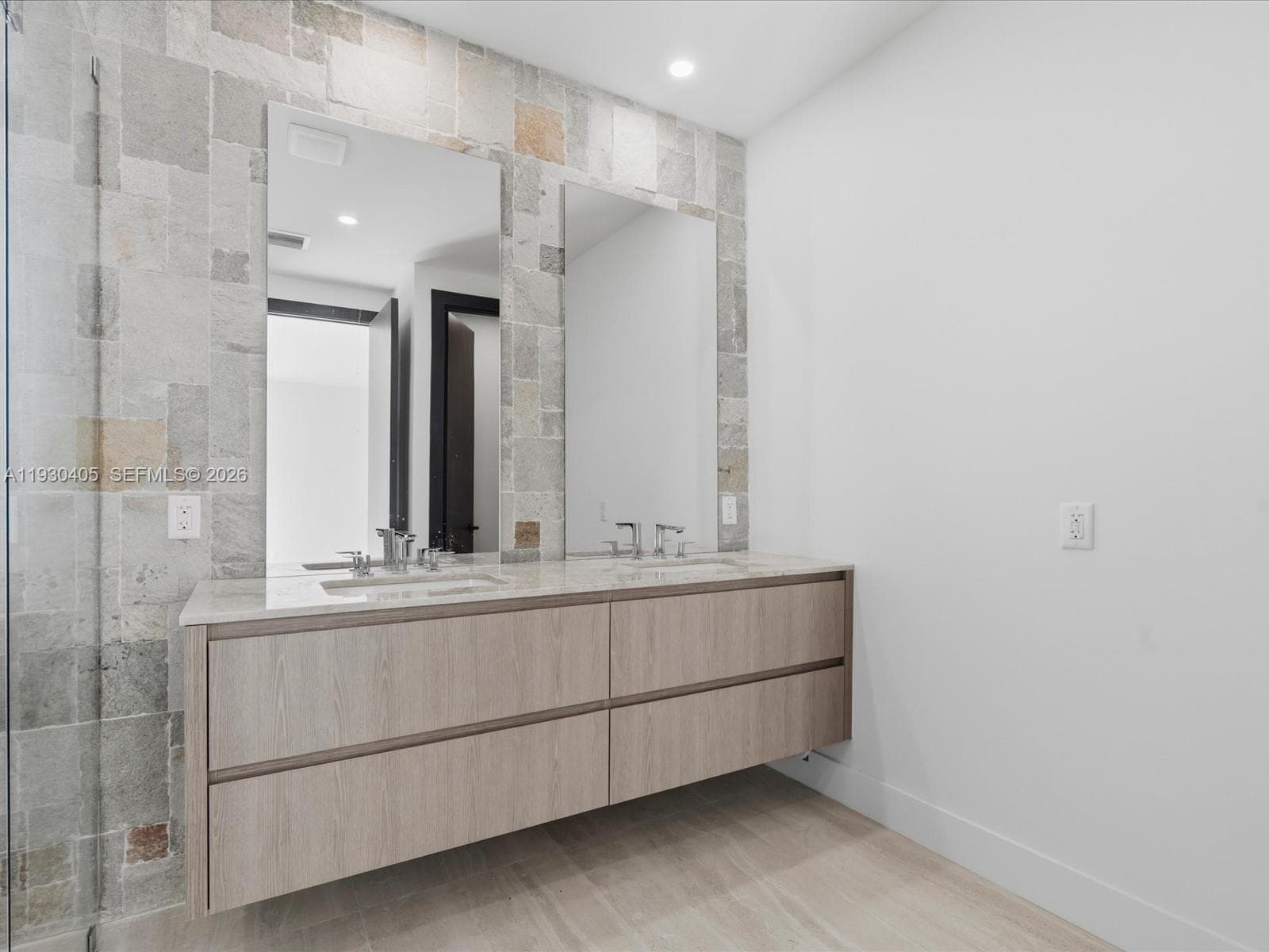 Floating vanity with wood-finish cabinetry, dual undermount sinks, stone-finish countertops, and natural stone tile backsplash with integrated vanity mirrors