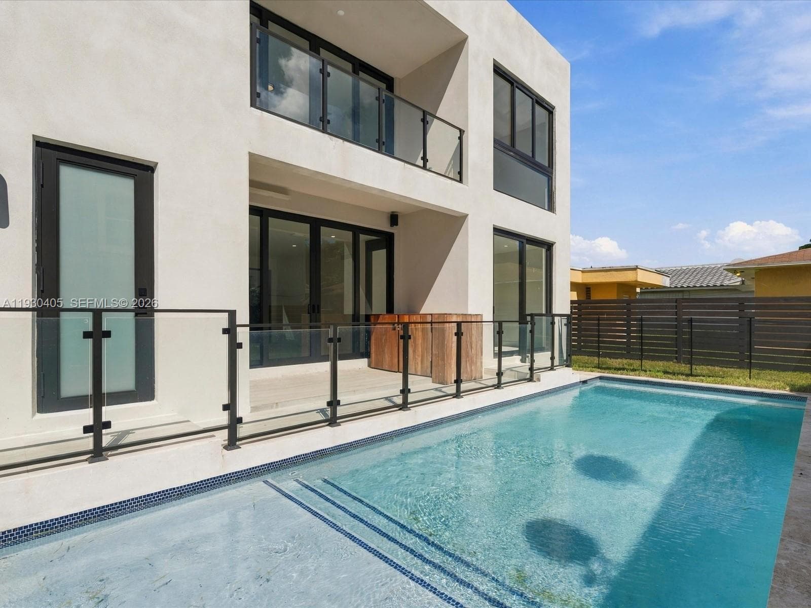 Modern two-story stucco exterior featuring a private swimming pool, glass balcony railings, expansive multi-panel sliding glass doors, and ground-level stone patio decking