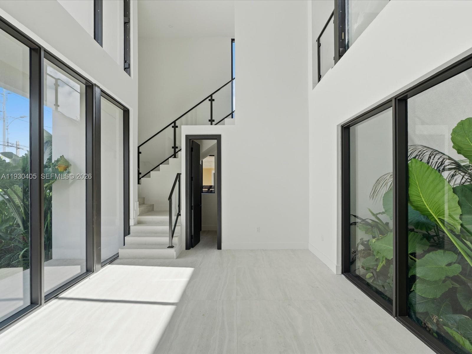 Double-height ceilings, floor-to-ceiling windows with black metal frames, stone-finish flooring, and a staircase featuring glass railing panels with black metal handrails
