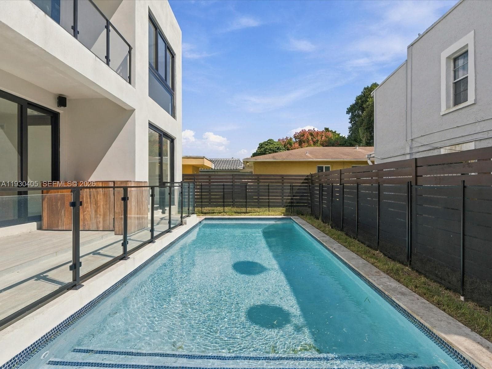 Rectangular in-ground pool with tiled waterline, concrete patio, glass-panel railing, and horizontal privacy fencing