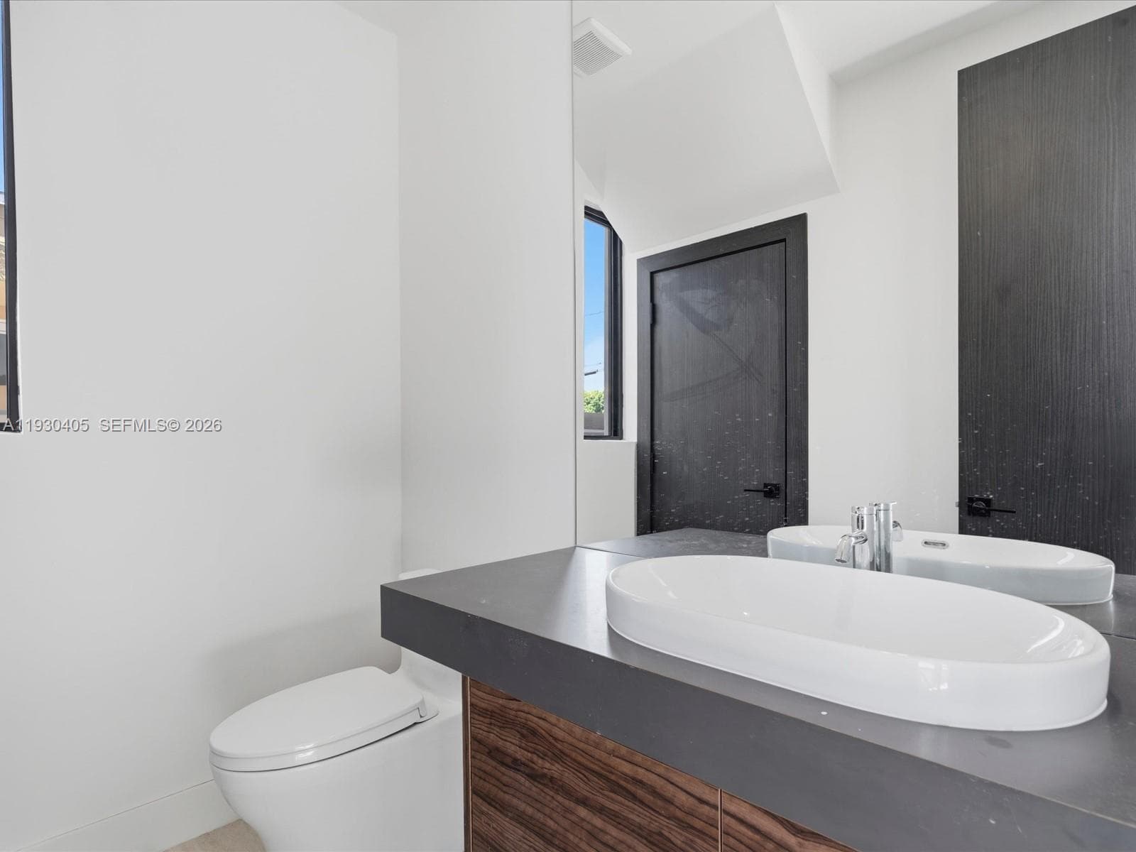 Modern floating vanity featuring a wood-finish base, dark countertop, and oval vessel sink, complemented by a large wall-mounted mirror, matte black hardware, and an elongated toilet