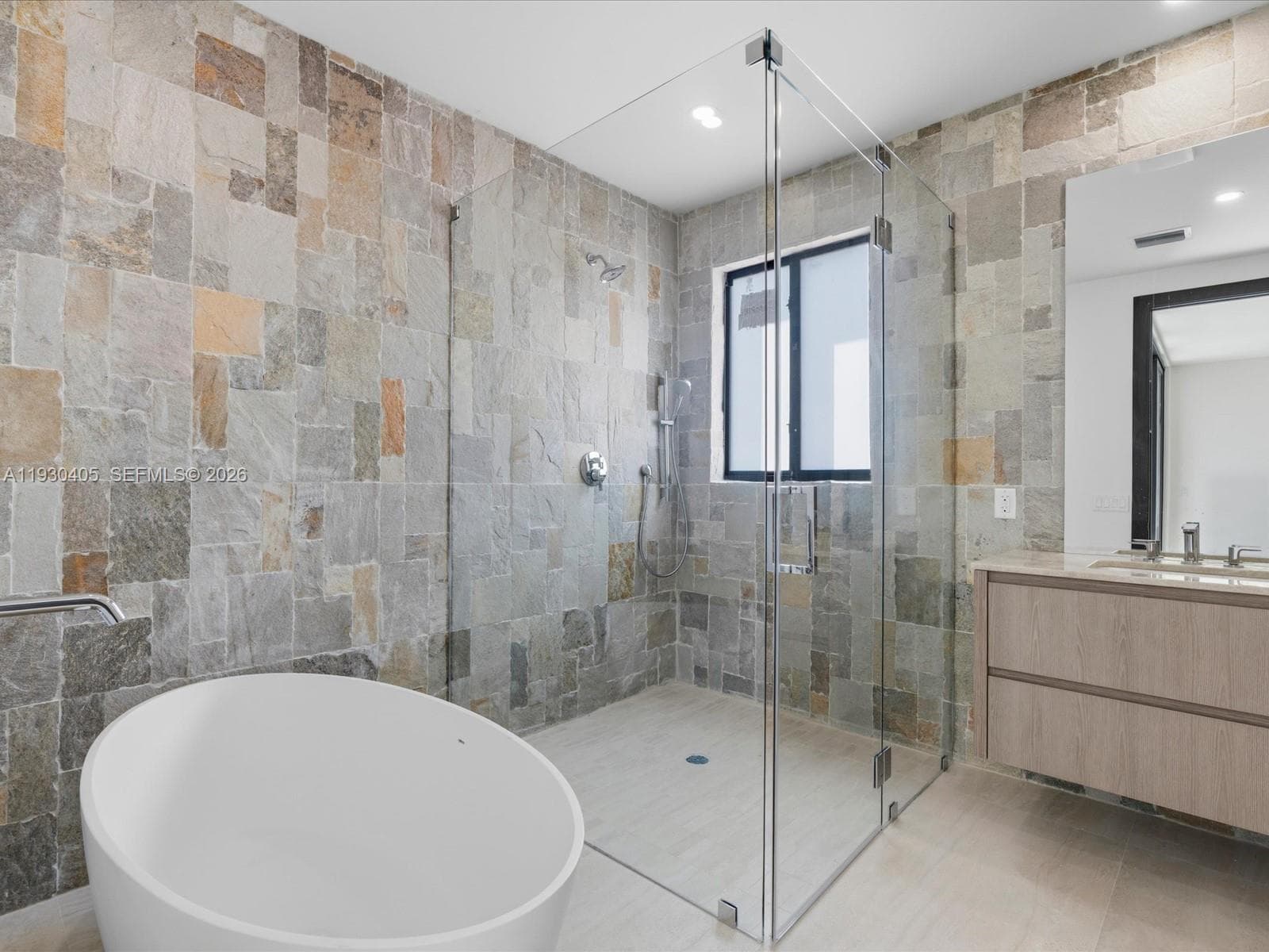 Freestanding soaking tub, frameless glass-enclosed shower, natural stone-tiled walls, wall-mounted floating vanity, and large-format tile flooring