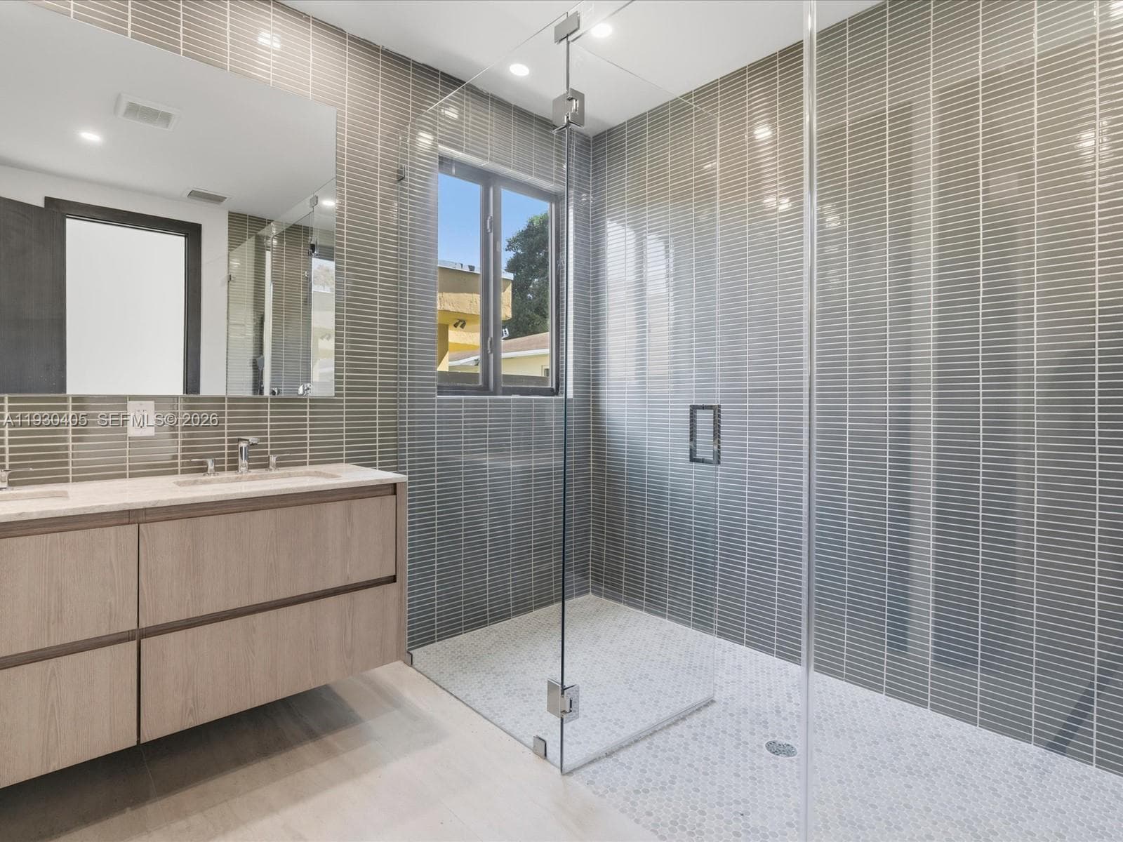 Floating vanity with light wood-finish cabinetry, quartz countertop, floor-to-ceiling vertically stacked tile, frameless glass shower enclosure, and circular mosaic floor tile