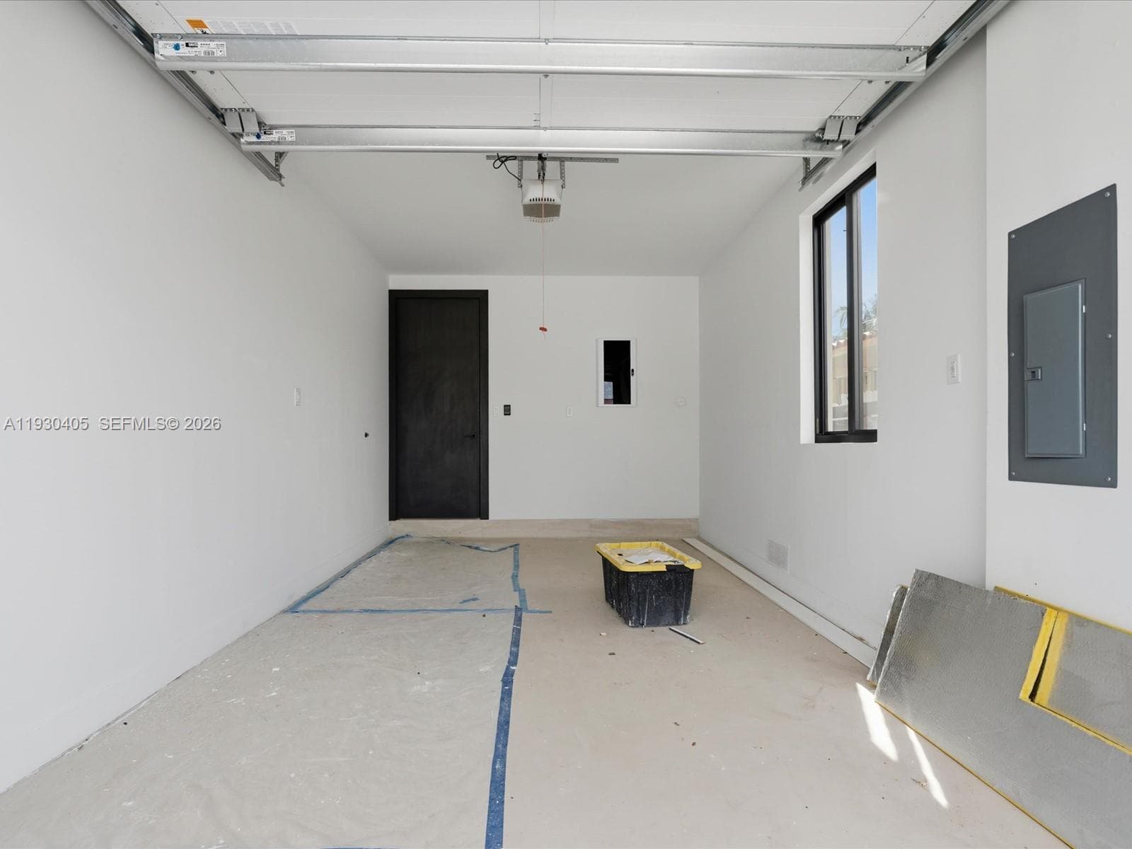 Finished garage interior featuring white-painted walls, a dark-toned interior door, a single-pane window with black trim, and an exposed ceiling-mounted garage door mechanism
