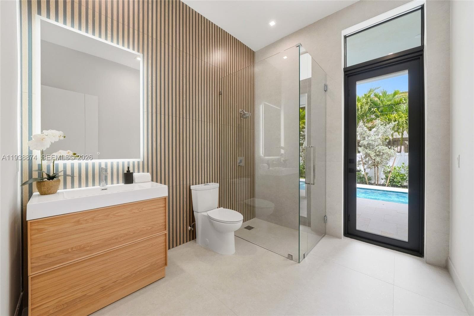 Bathroom featuring a wood-finish floating vanity, wall-mounted illuminated mirror, vertical slat-wood accent wall, glass-enclosed shower, and exterior door access