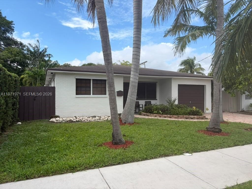 Painted brick exterior featuring a recessed entryway, attached garage with roll-up door, horizontal window shutters, and landscaped front lawn with palm trees