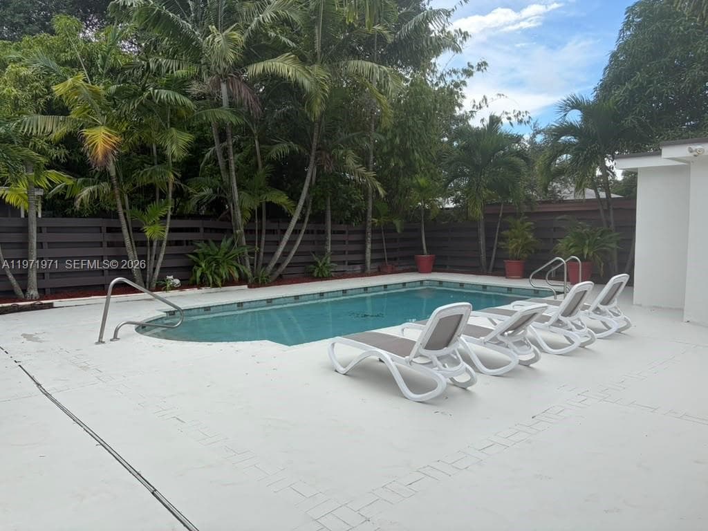 In-ground swimming pool with metal handrails, surrounded by expansive concrete decking and a perimeter privacy fence, accented by mature palm trees