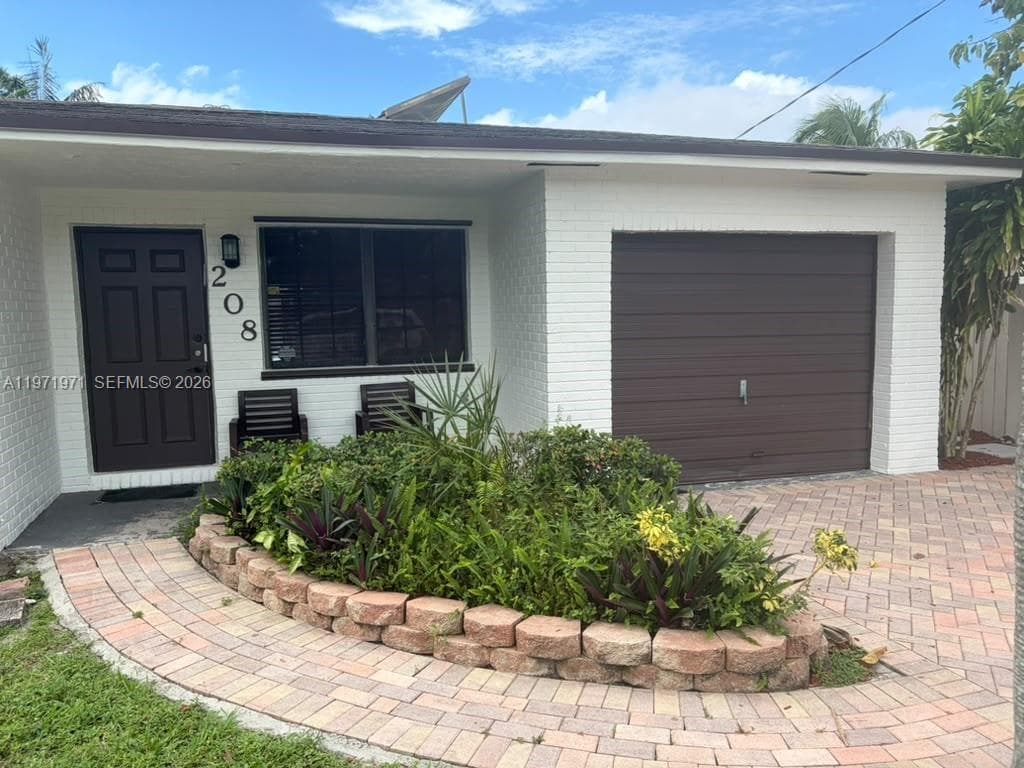 White-painted brick exterior featuring an integrated garage, dark-toned entry door, brick-paved driveway, and a raised stone-block landscape bed