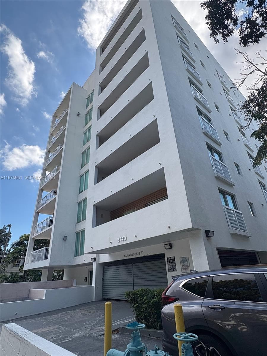 Mid-rise stucco exterior featuring stacked recessed balconies, floor-to-ceiling glass windows, and a secure gated garage entrance