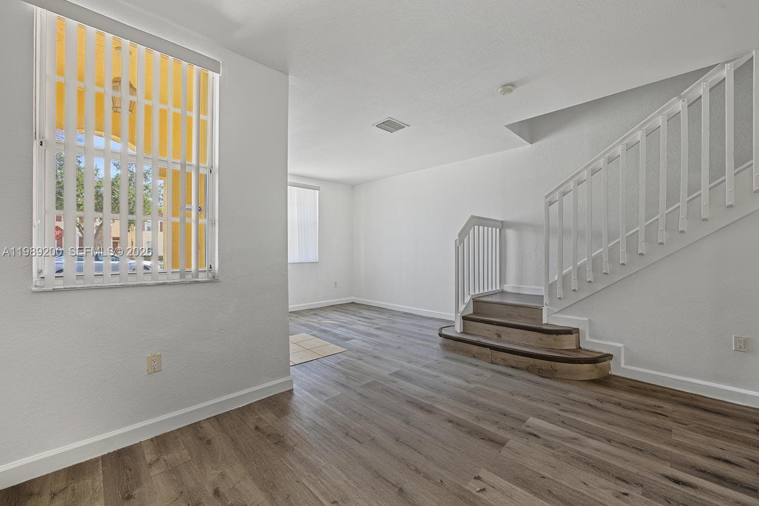 Interior space featuring wood-finish flooring, a staircase with decorative curved bottom treads, white baluster railings, and windows with vertical blinds