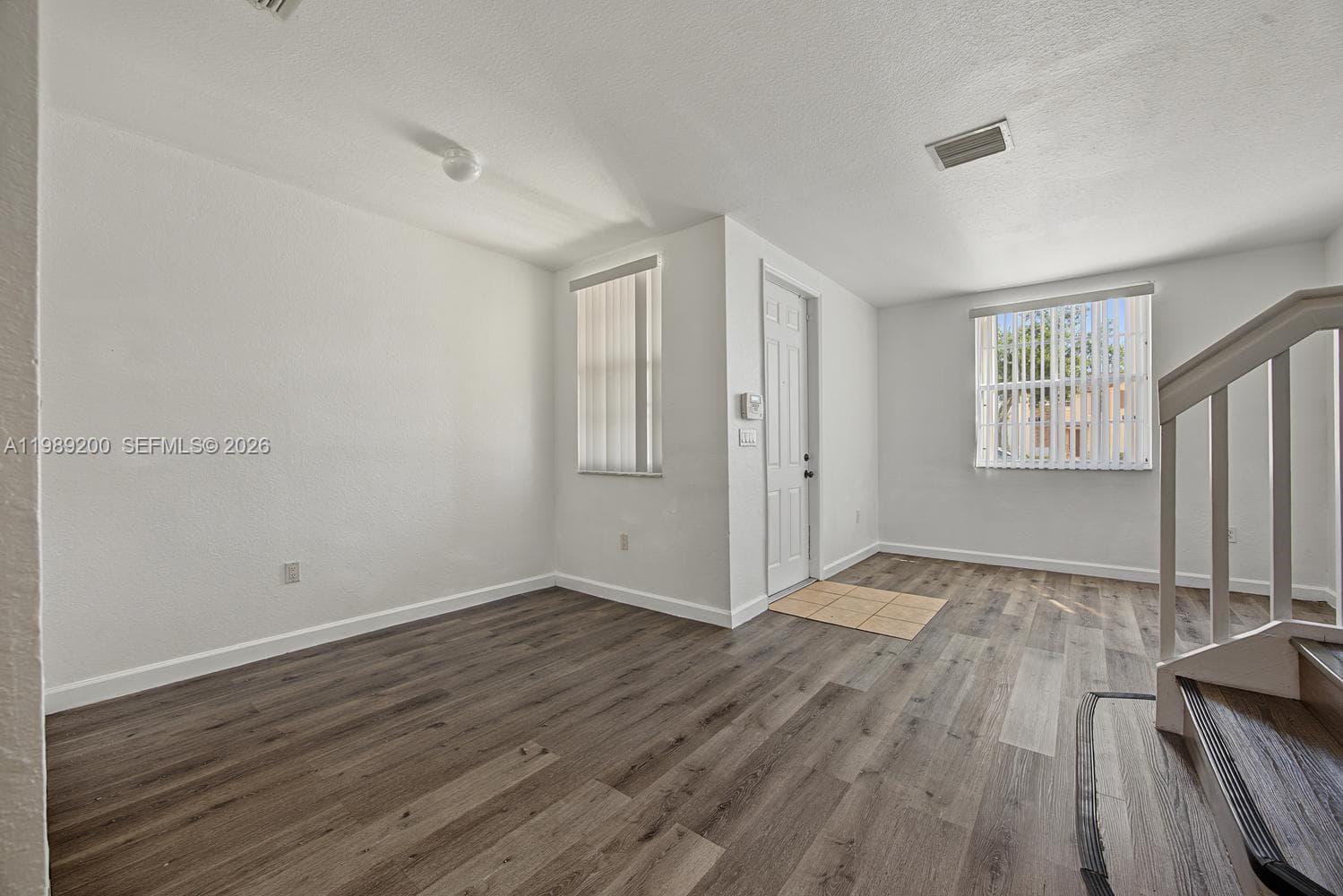 Wood-finish flooring, neutral walls, two windows with vertical blinds, tile-floored entry, and staircase with wooden handrail