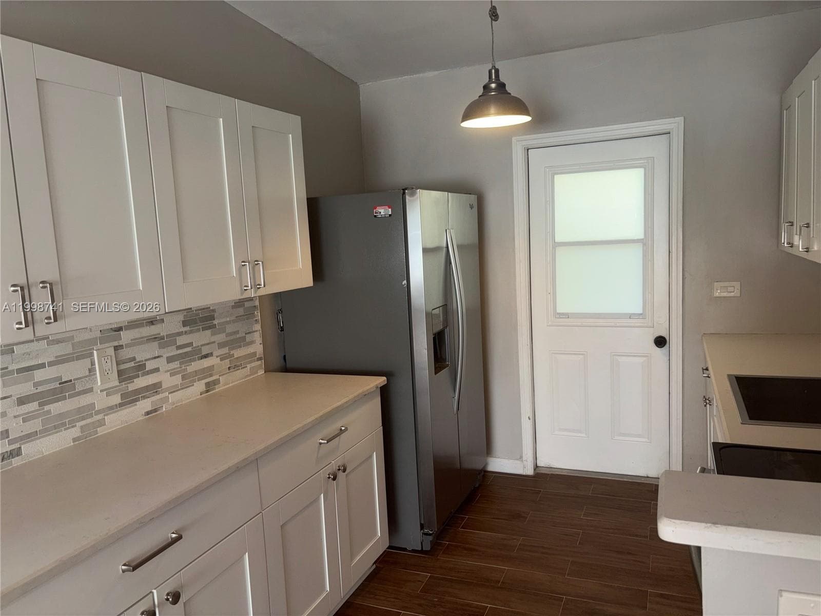 Kitchen featuring white shaker-style cabinetry, multi-toned linear tile backsplash, stainless steel refrigerator, wood-finish flooring, and a single pendant light fixture