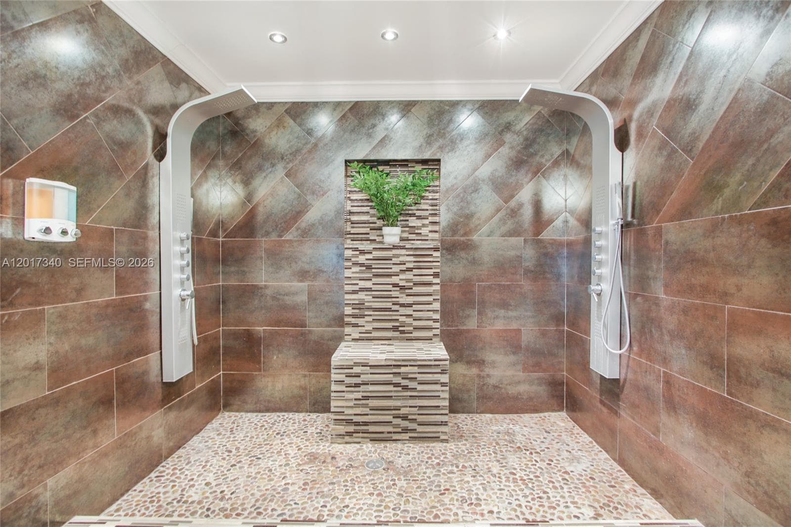 Walk-in shower featuring large-format metallic-finish wall tiles, dual vertical shower panels with integrated body sprayers, pebble-stone flooring, and a built-in tiled bench with a recessed decorative accent wall