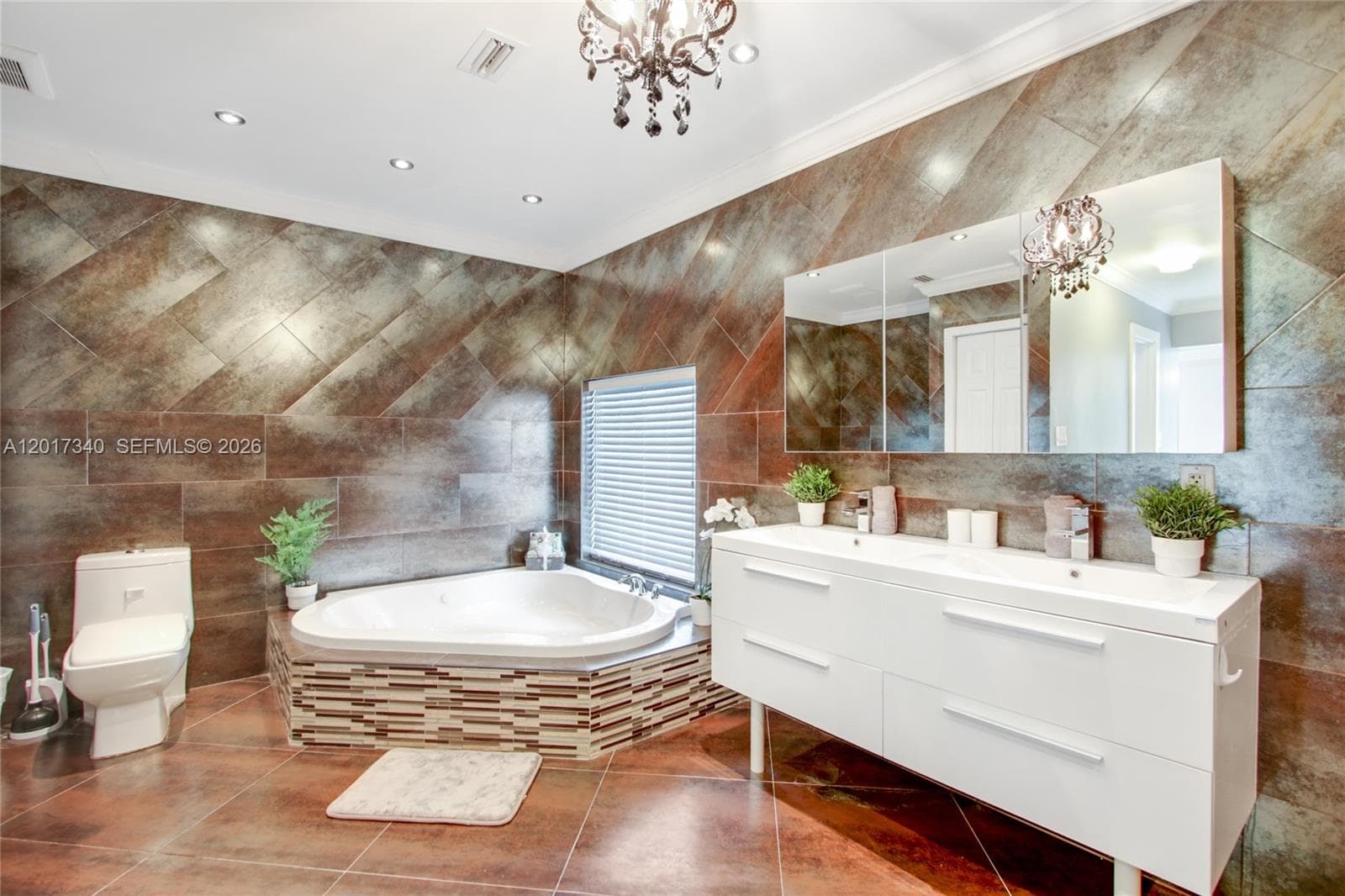 Bathroom featuring large-format diagonal tile wall cladding, a corner jetted soaking tub with mosaic tile surround, a white double-vanity cabinet with integrated sinks, and ornate chandelier lighting