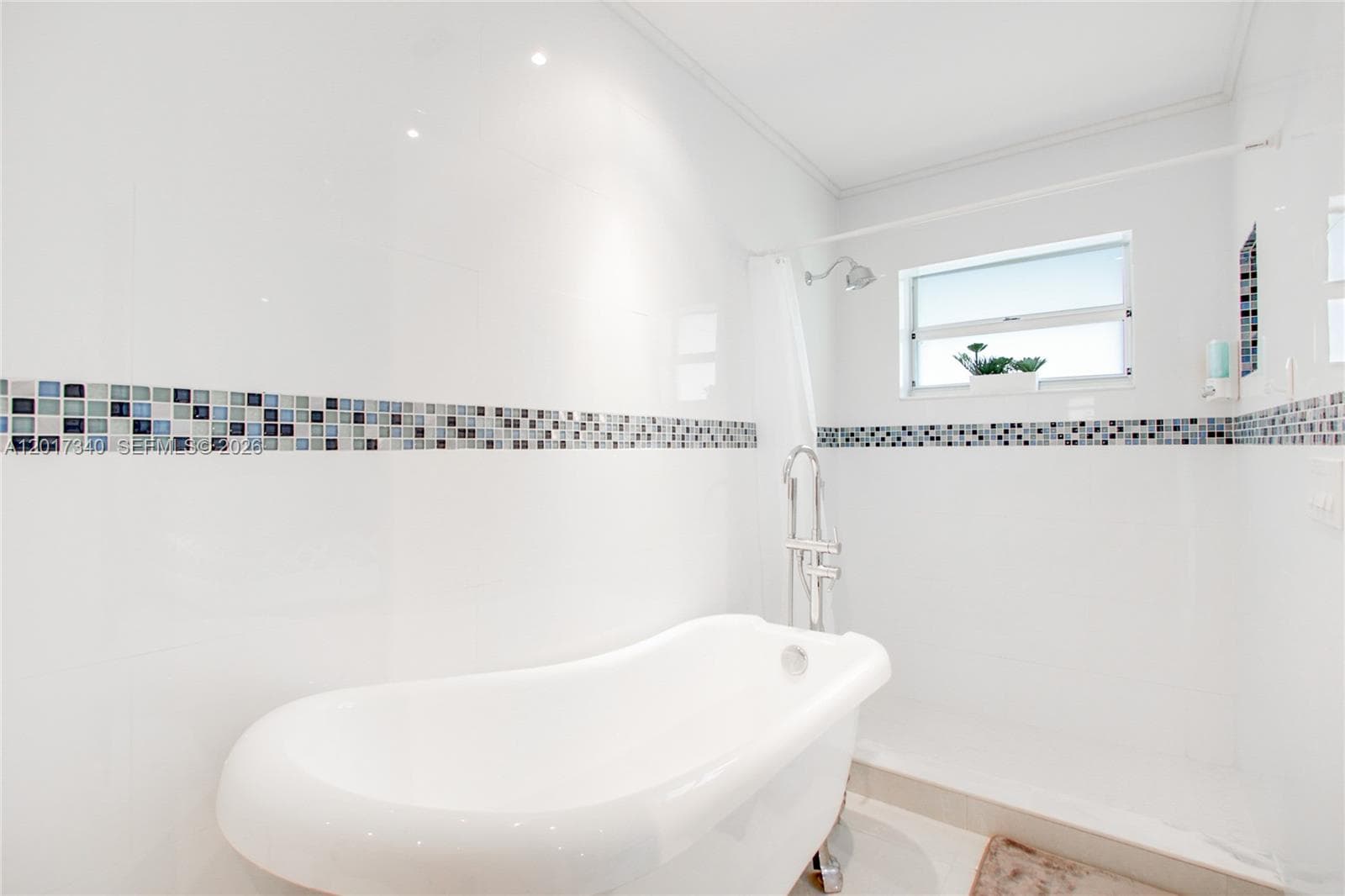 Freestanding soaking tub with a chrome floor-mounted faucet, crisp white wall tile, decorative mosaic tile inlay, and recessed lighting