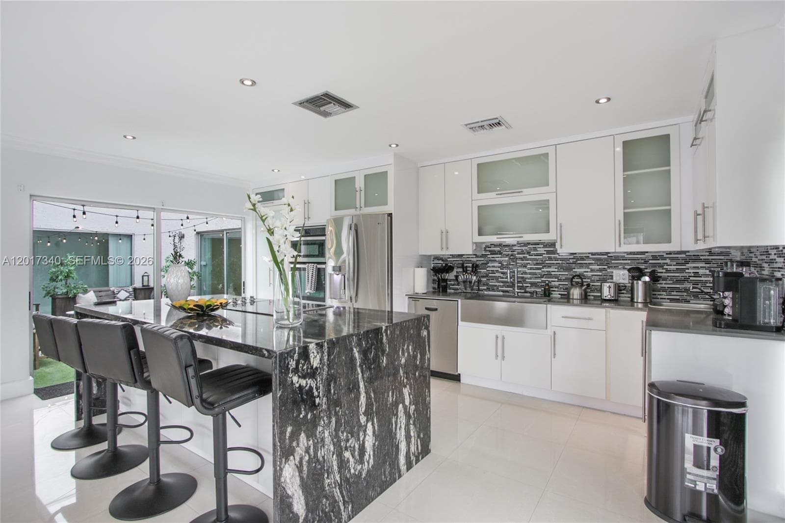 Kitchen featuring white cabinetry with glass-panel accents, large center island with dark natural stone countertop, apron-front stainless steel sink, tiled backsplash, and recessed ceiling lighting