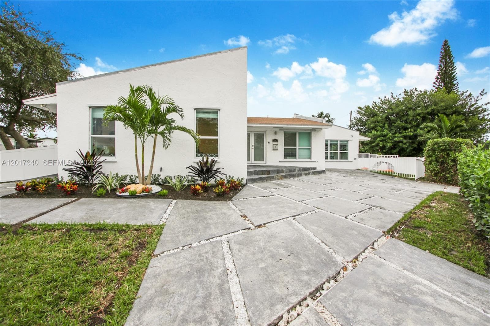 Mid-century modern exterior featuring a distinct slanted roofline, white stucco facade, large-format concrete paver driveway with decorative stone inlay, and manicured landscaping