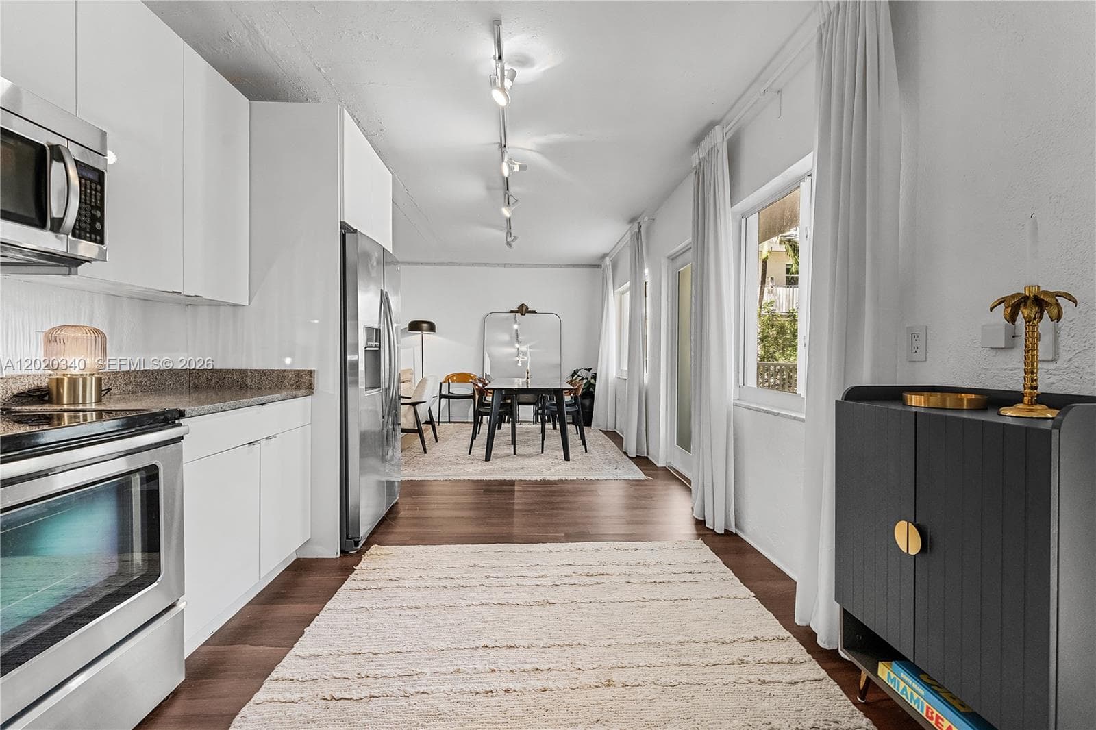 A modern open-plan kitchen and dining area with white cabinetry, stainless steel appliances, dark hardwood floors, and a cream runner rug. Warm gold accents — including a palm tree sculpture and brass lamp — give it a Miami-inspired flair. Bright and airy with natural light from sheer-curtained windows.