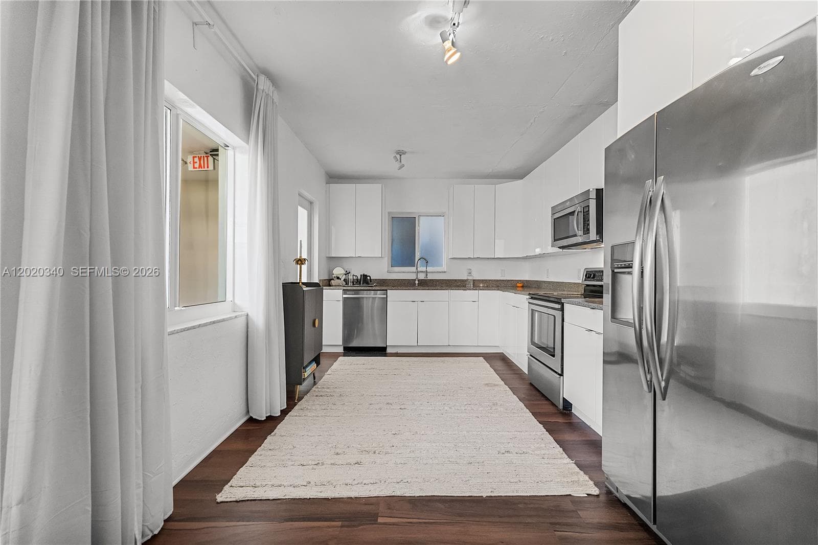 A clean, U-shaped kitchen with white flat-front cabinets, dark granite countertops, and stainless steel appliances, set on rich dark hardwood floors with sheer white curtains and track lighting.