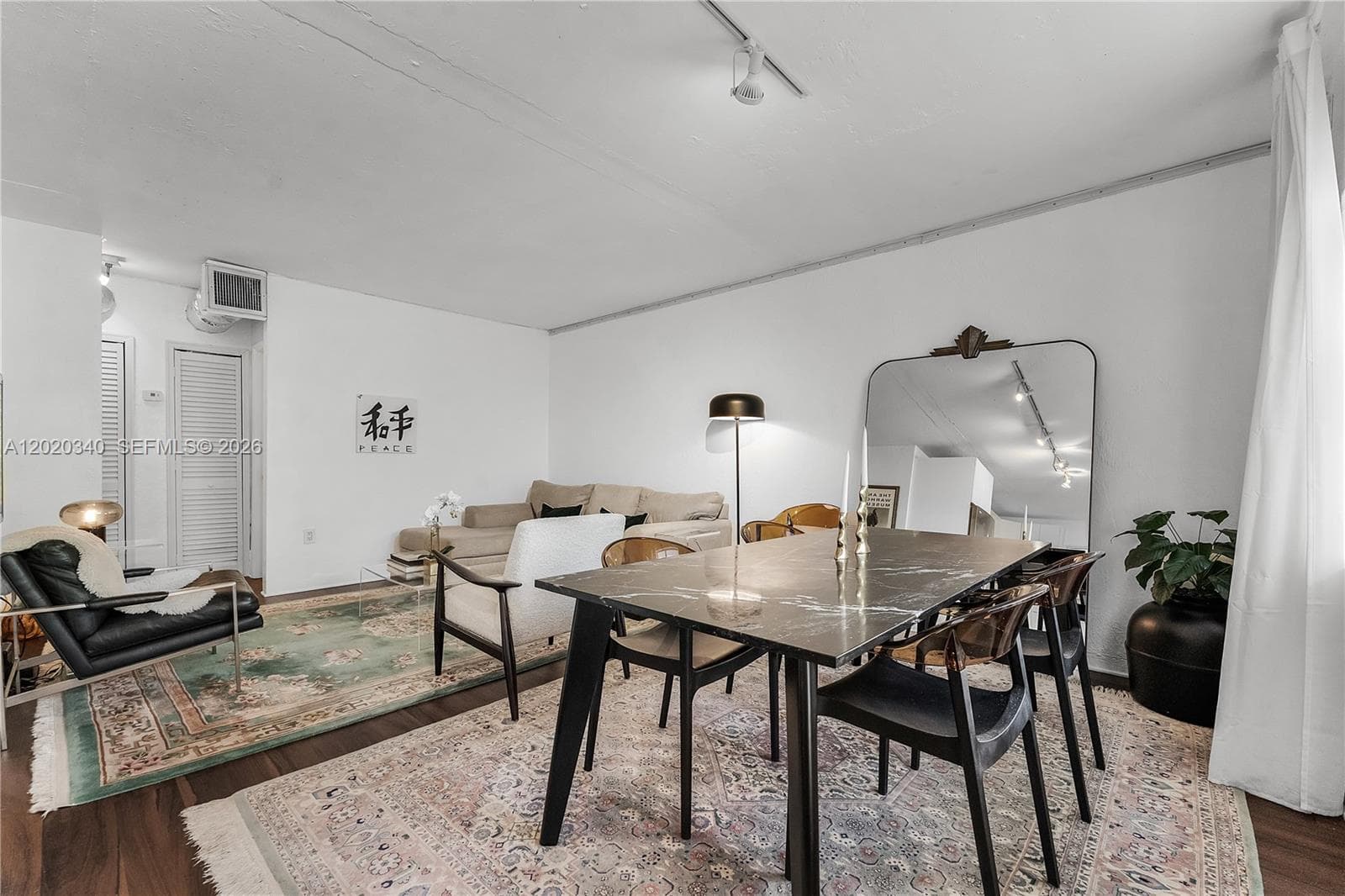 An eclectic open-plan dining and living space with a dark marble-top dining table, mixed wood and black chairs, and a large leaning mirror. A beige sectional and vintage-style rugs anchor the living area, with a "Peace" kanji print and black leather accent chair adding personality. Warm and curated.