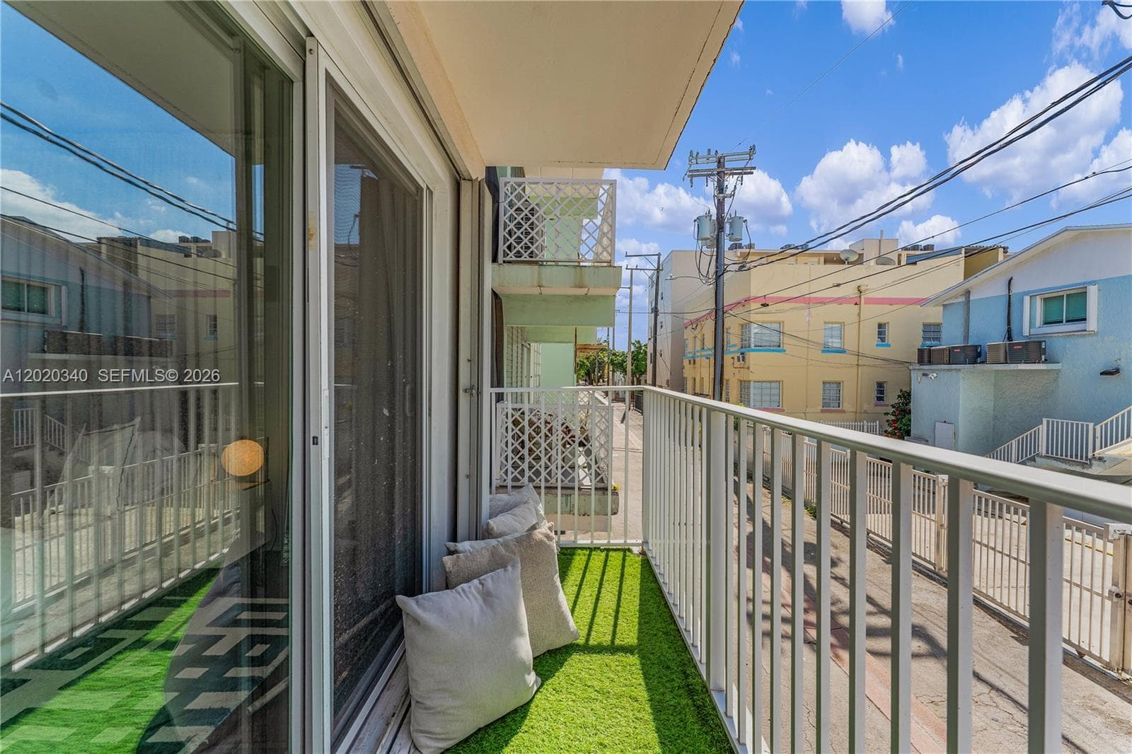 A narrow balcony laid with artificial turf and styled with a few scatter cushions for casual outdoor lounging. A sliding glass door connects it to the interior, reflecting the bright surroundings. The white metal railing overlooks a lively neighborhood streetscape of pastel-colored mid-rise buildings — mint green, yellow, and blue — under a sunny, partly cloudy sky typical of South Florida.